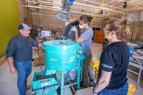 Three people pour dry concrete into a mixer inside a studio in the Iowa State University Communications Building.