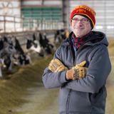 Matt McGarry in a coat, hat and gloves in a cattle barn, with dairy cows visible in the background.