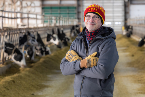 Matt McGarry in a coat, hat and gloves in a cattle barn, with dairy cows visible in the background.